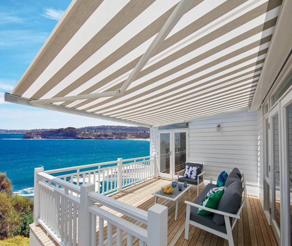 A beautiful outdoor view with a minimalist white long bench, chair, table, under a Luxaflex® Folding Arm Retractable Awnings assembled by Katandra Interiors Busselton.